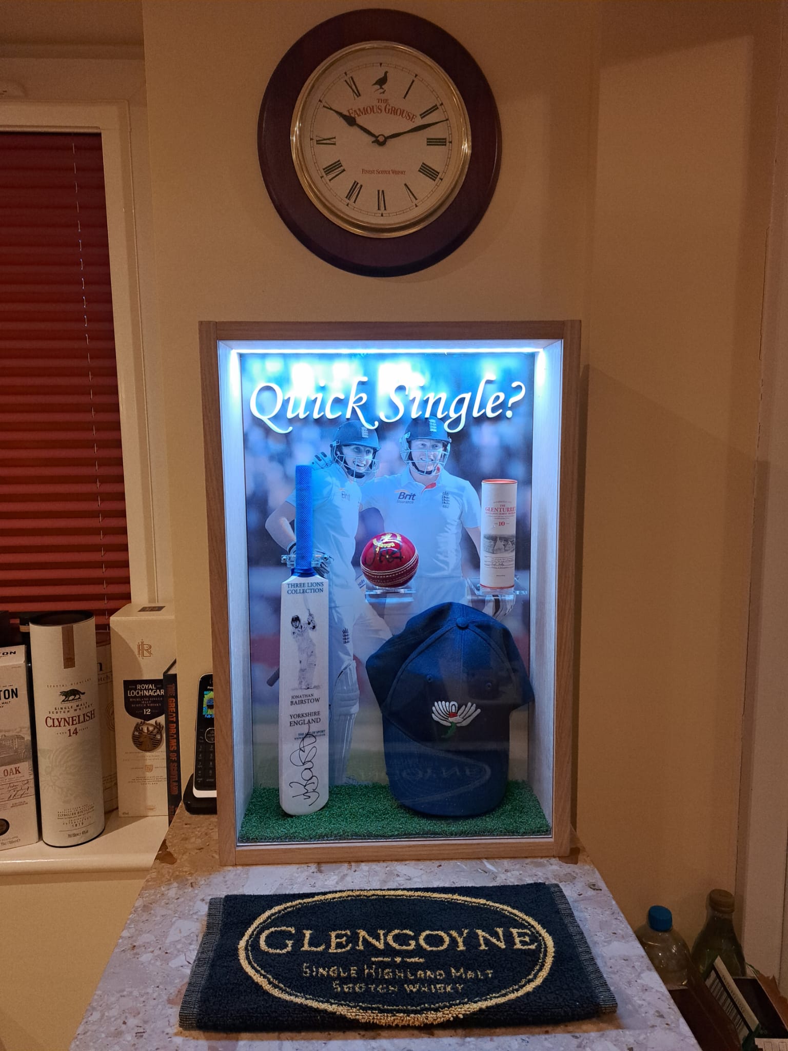 Illuminated acrylic display cabinet housing a signed cricket bat, red ball, cap and whisky bottle, with a Famous Grouse clock above