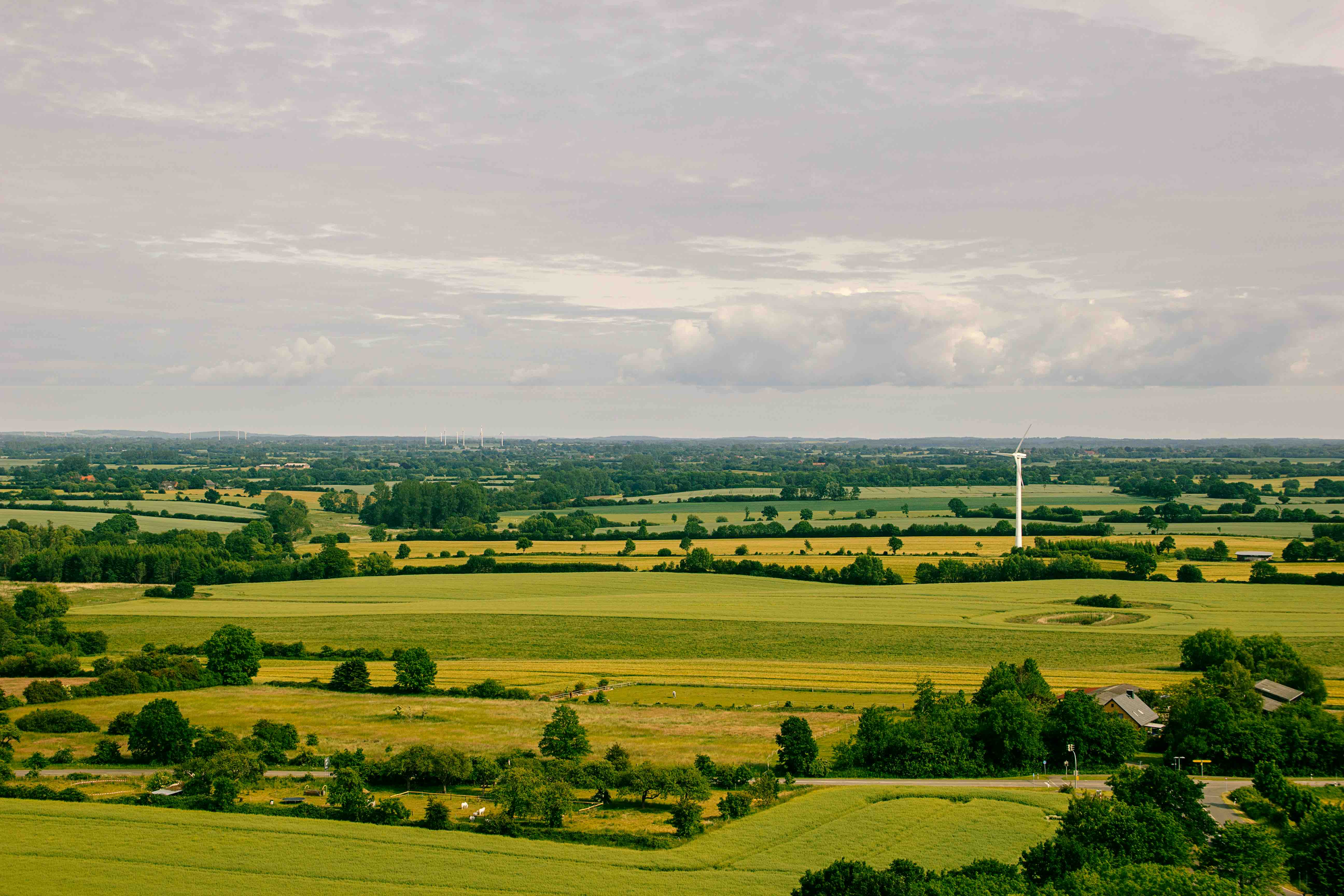 Aerial view of a vast rural landscape with green and golden fields, scattered trees, a wind turbine, and overcast skies