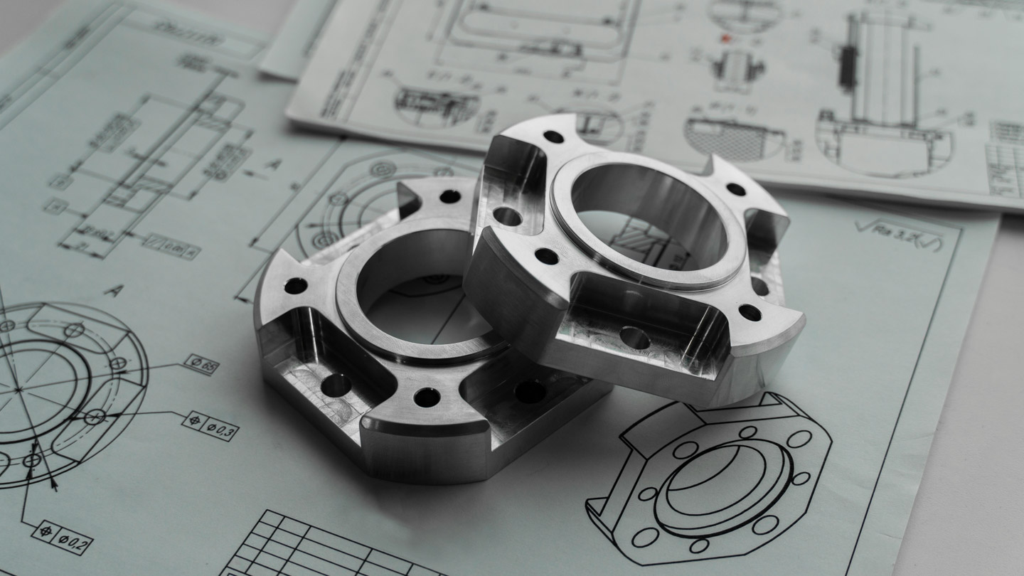 Two precision-machined metal flanged components resting on detailed engineering technical drawings