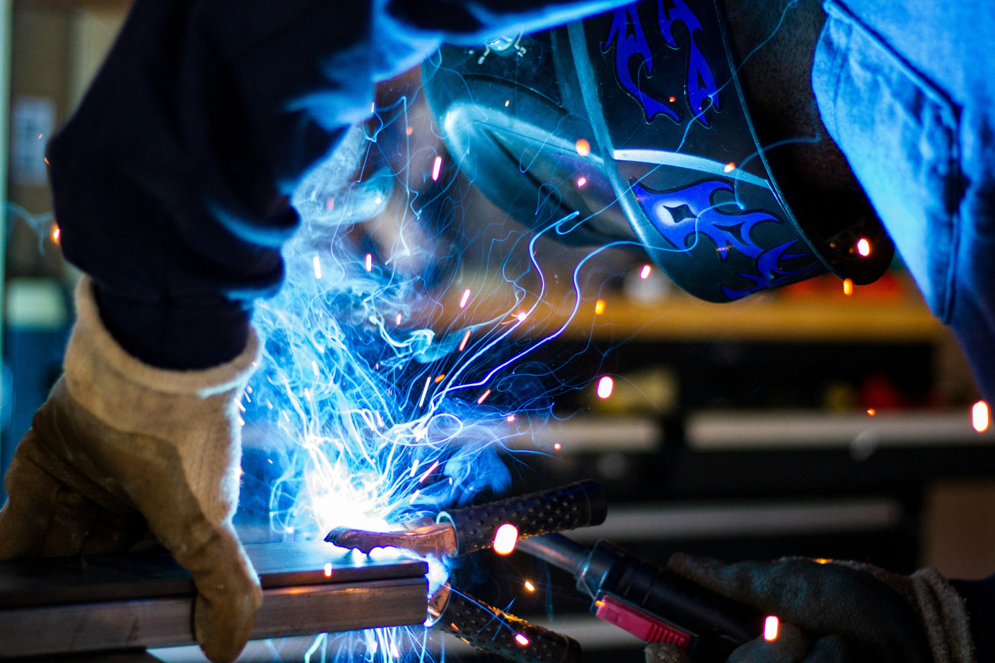Welder in a flame-decorated helmet and leather gloves MIG welding metal, with bright blue sparks and smoke flying