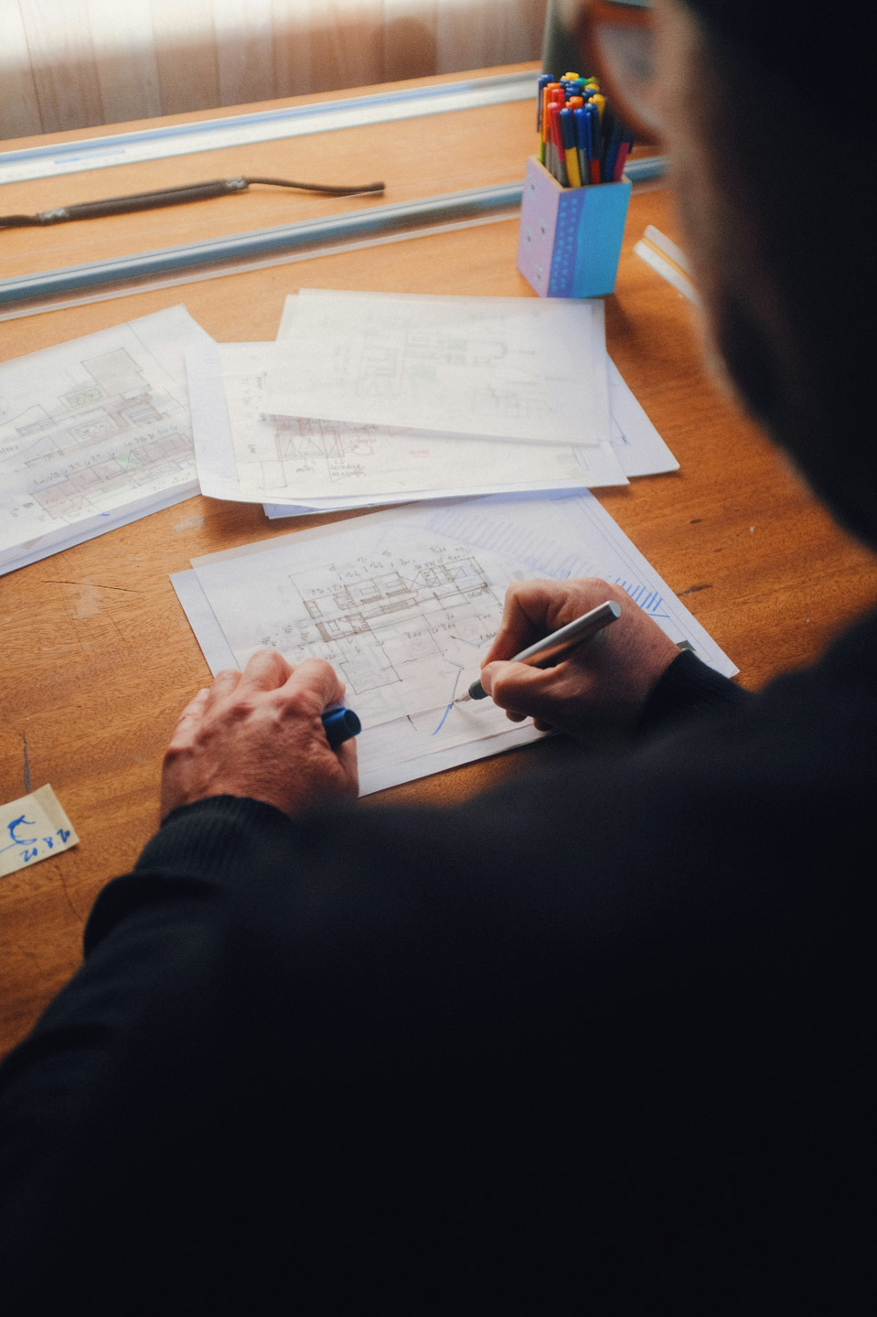 A person drawing architectural floor plan sketches at a wooden desk with scattered papers and a pot of coloured pens nearby
