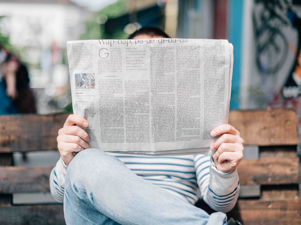 Person seated outdoors on a wooden bench, holding up a newspaper that obscures their face, wearing a striped top and jeans