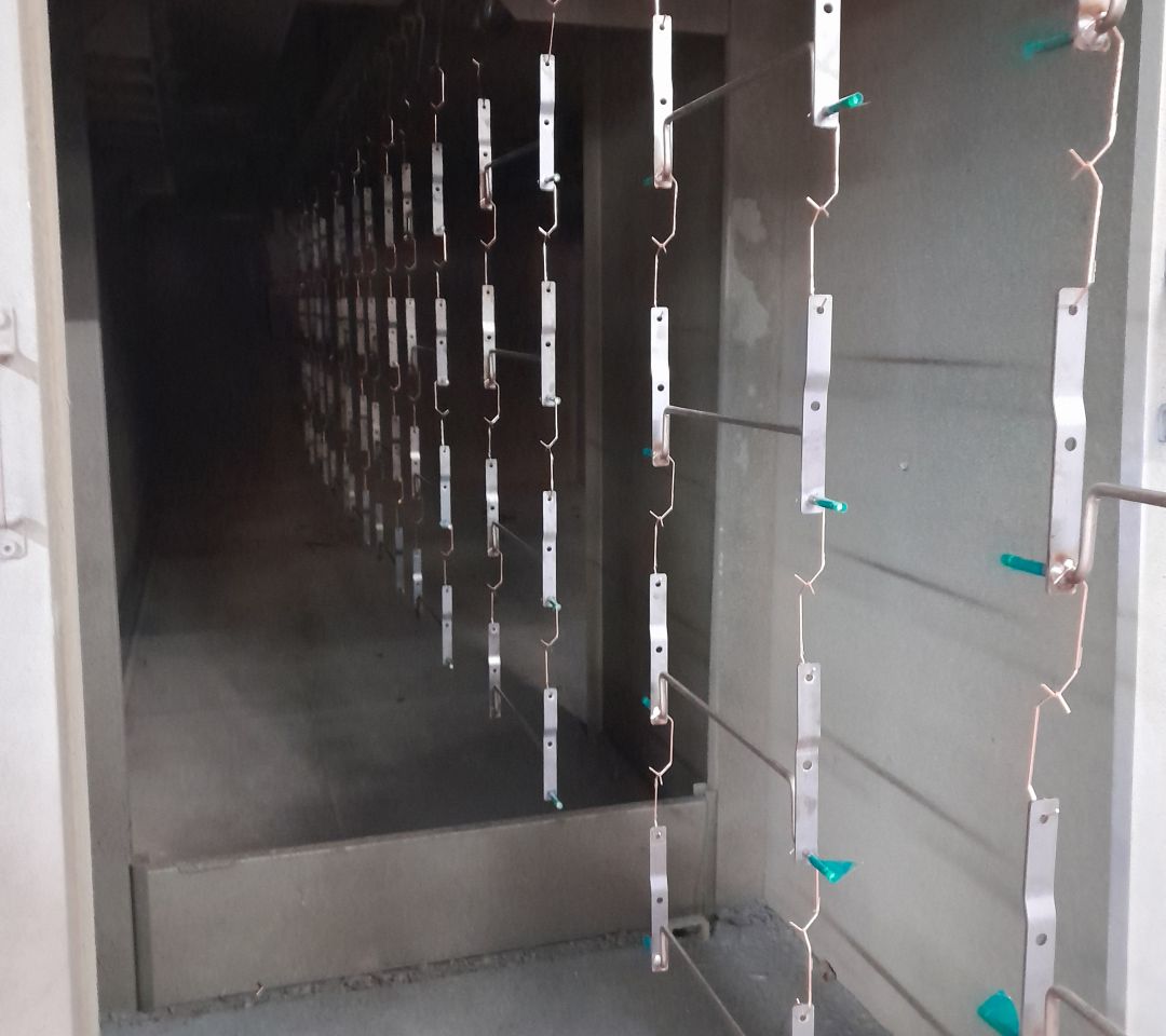 Metal brackets suspended on hanging racks inside an industrial powder coating oven tunnel with teal plastic hooks