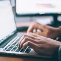 Person typing on a silver laptop at a desk, with a second monitor visible in the background