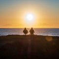 Two silhouetted figures seated in chairs on a clifftop, watching a golden sunset over the ocean.
