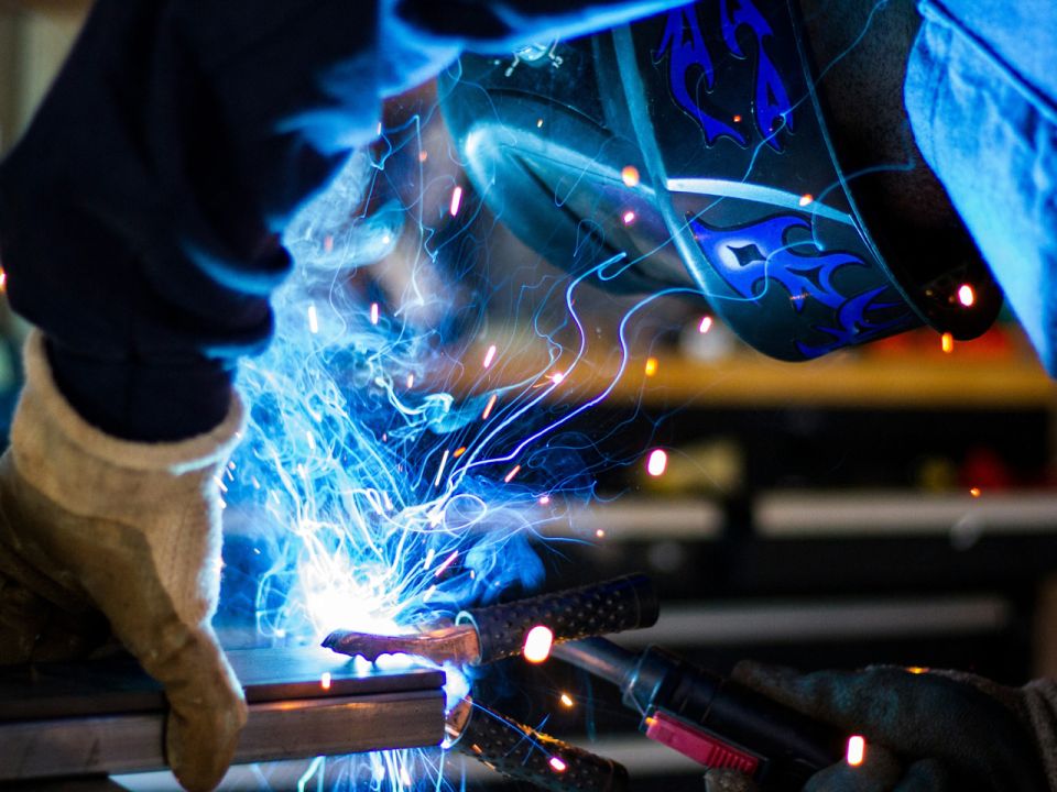 Welder in a flame-decorated helmet and leather gloves MIG welding metal, with bright blue sparks and smoke flying