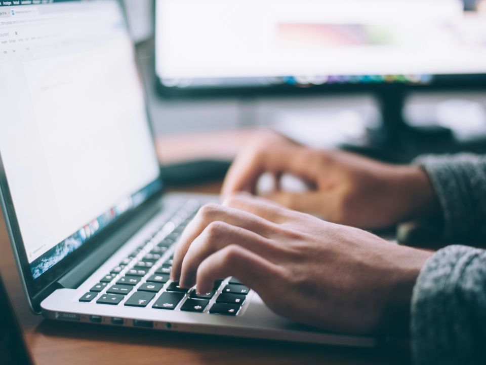Person typing on a silver laptop at a desk, with a second monitor visible in the background