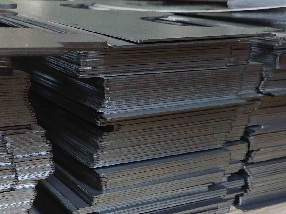 Large stack of dark sheet metal plates piled on a ridged concrete floor in an industrial setting