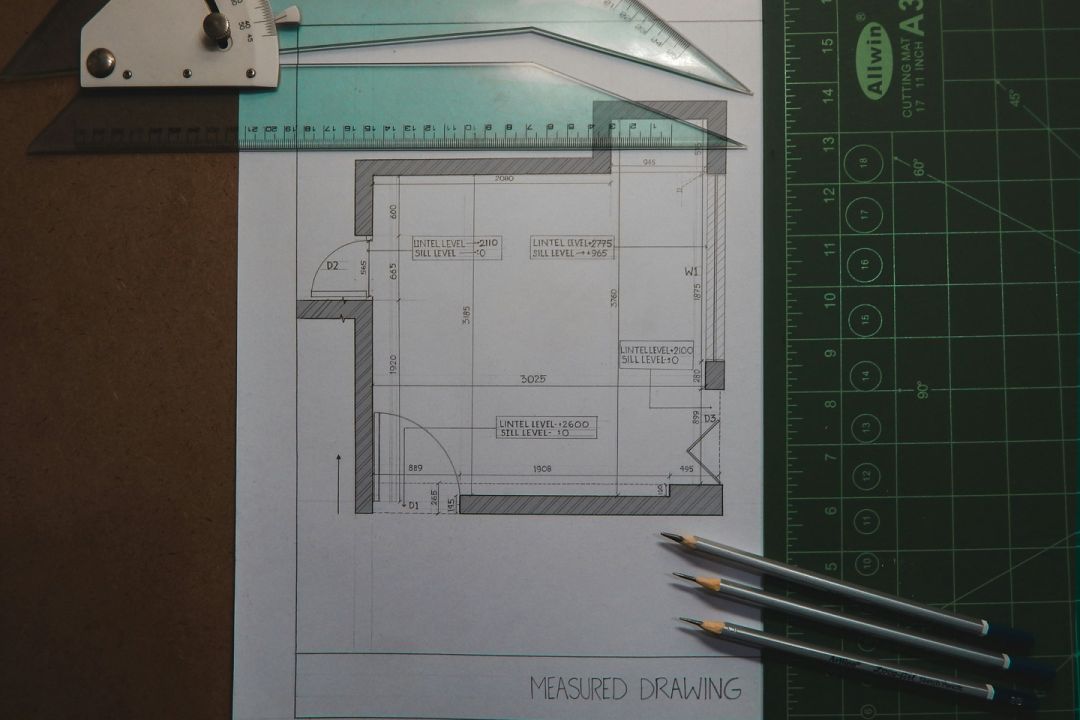 Hand-drawn architectural measured drawing on paper, surrounded by rulers, set squares, and pencils on a cutting mat