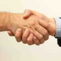 Two people shaking hands, one wearing a navy suit jacket with a blue checked shirt cuff visible