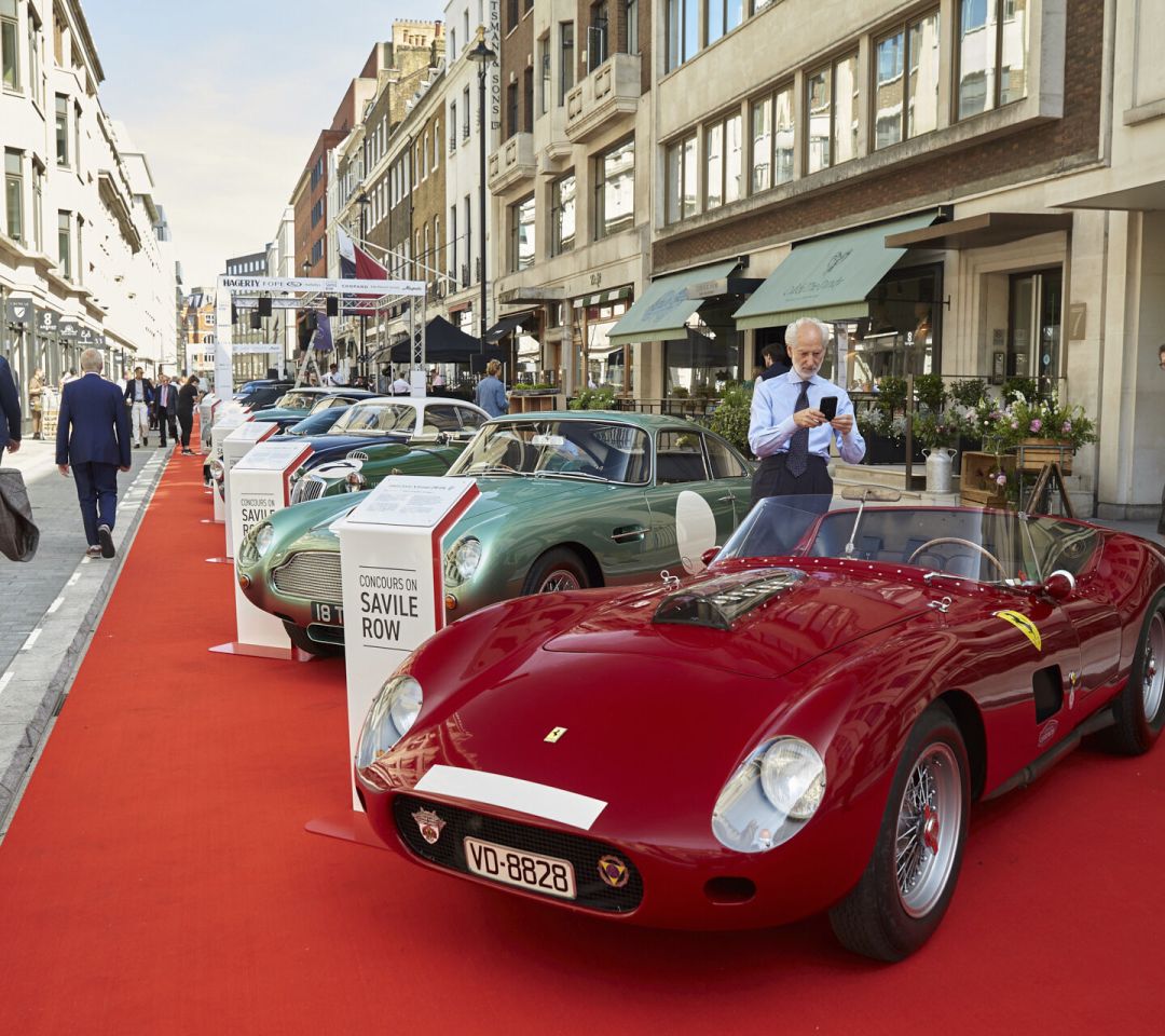 Classic cars including a red Ferrari and green Aston Martin displayed on a red carpet at the Concours on Savile Row event.