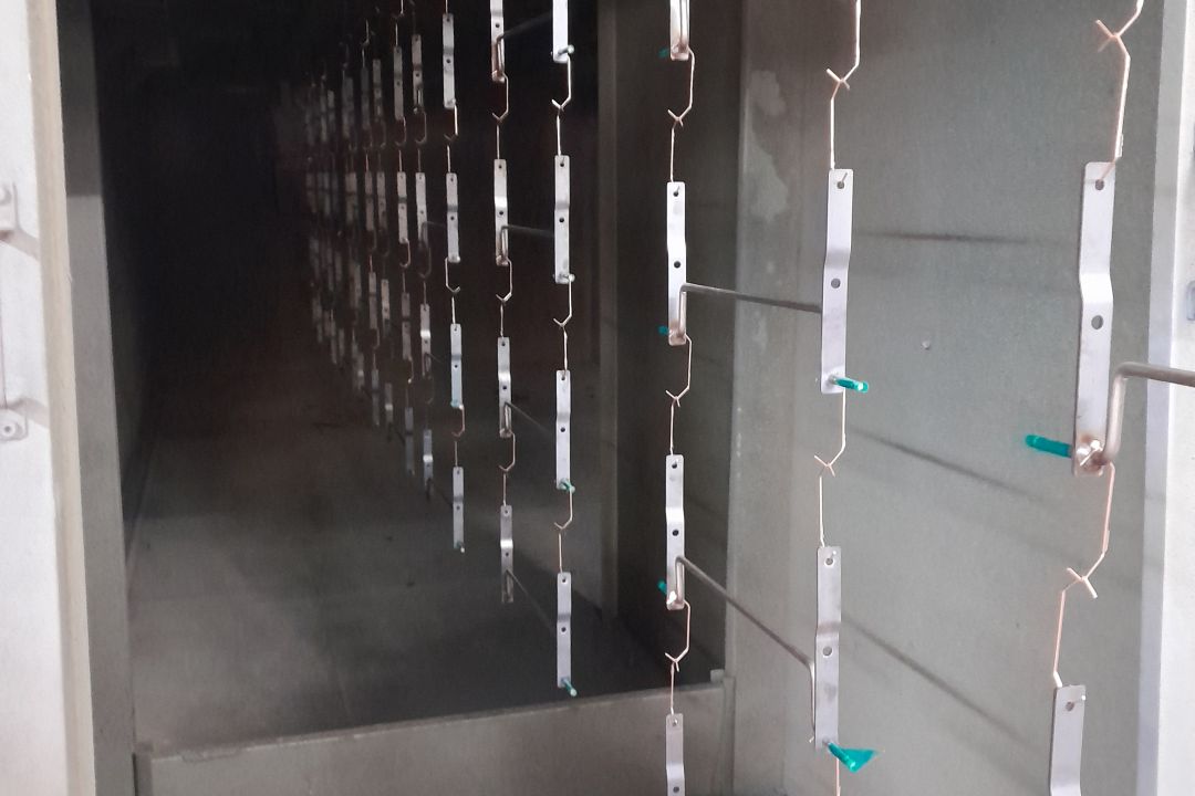 Metal brackets suspended on hanging racks inside an industrial powder coating oven tunnel with teal plastic hooks