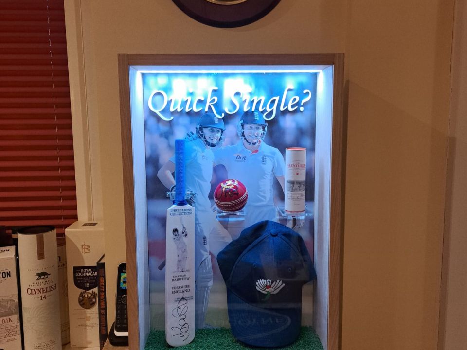 Illuminated acrylic display cabinet housing a signed cricket bat, red ball, cap and whisky bottle, with a Famous Grouse clock above