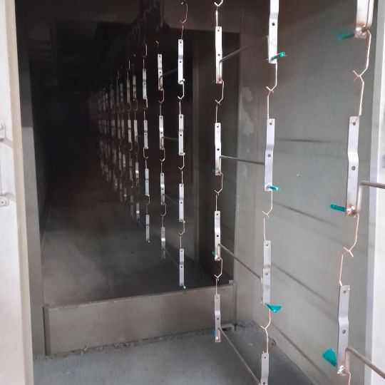 Metal brackets hanging on chains inside an industrial powder coating oven tunnel with green plastic clips visible
