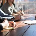 A group of people seated at a wooden table during a meeting, taking notes in notebooks with pens