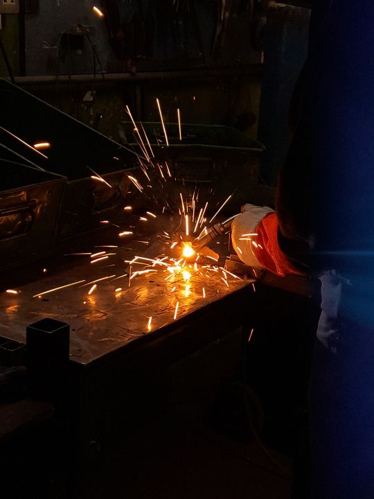 A welder's gloved hand welding steel plate in a dark workshop, sending bright orange sparks flying across the metal surface.