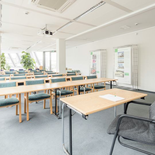 Bright classroom layout with rows of light wood tables, teal padded chairs, and a ceiling-mounted projector