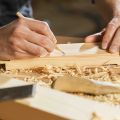A carpenter marking a measurement on a timber plank with a pencil, surrounded by wood shavings on a workbench