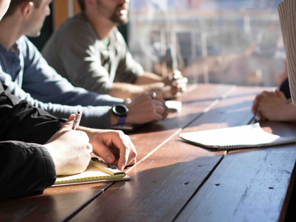 A group of people seated at a wooden table during a meeting, taking notes in notebooks with pens