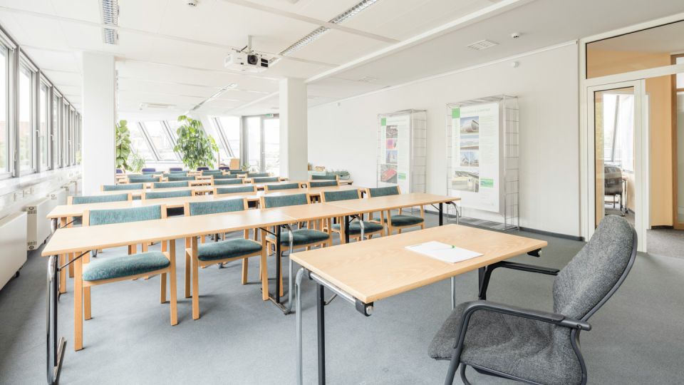 Bright classroom layout with rows of light wood tables, teal padded chairs, and a ceiling-mounted projector