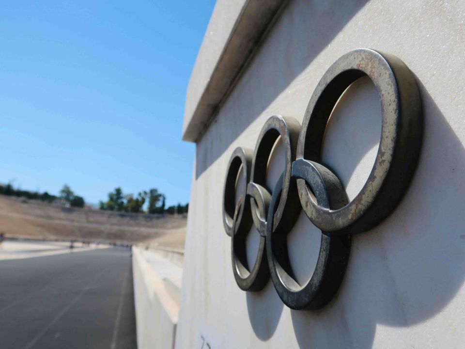 Close-up of a weathered metal Olympic rings emblem mounted on a white stone wall at an empty stadium
