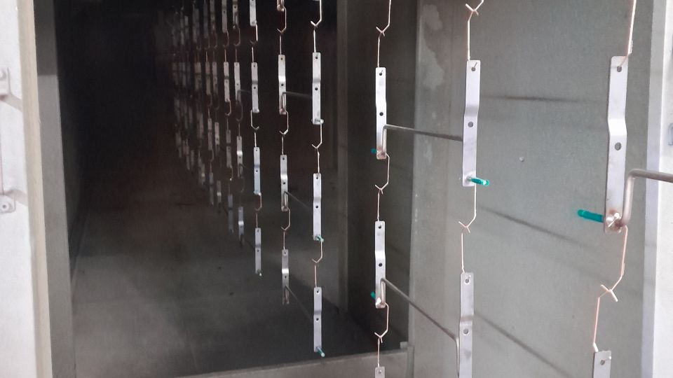 Metal brackets hanging on chains inside an industrial powder coating oven tunnel with green plastic clips visible