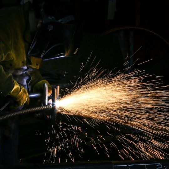 A welder in a protective mask and yellow gloves welding metal, sending a bright cascade of sparks across a dark workshop.