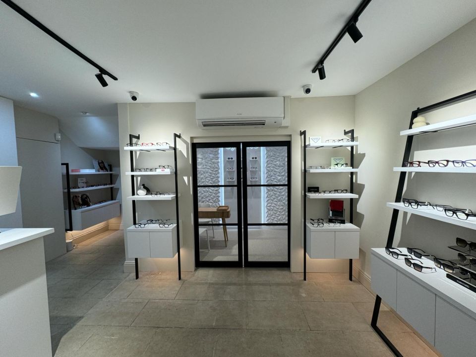 Interior of an optician's showroom with white shelving units displaying glasses frames and sunglasses on both walls