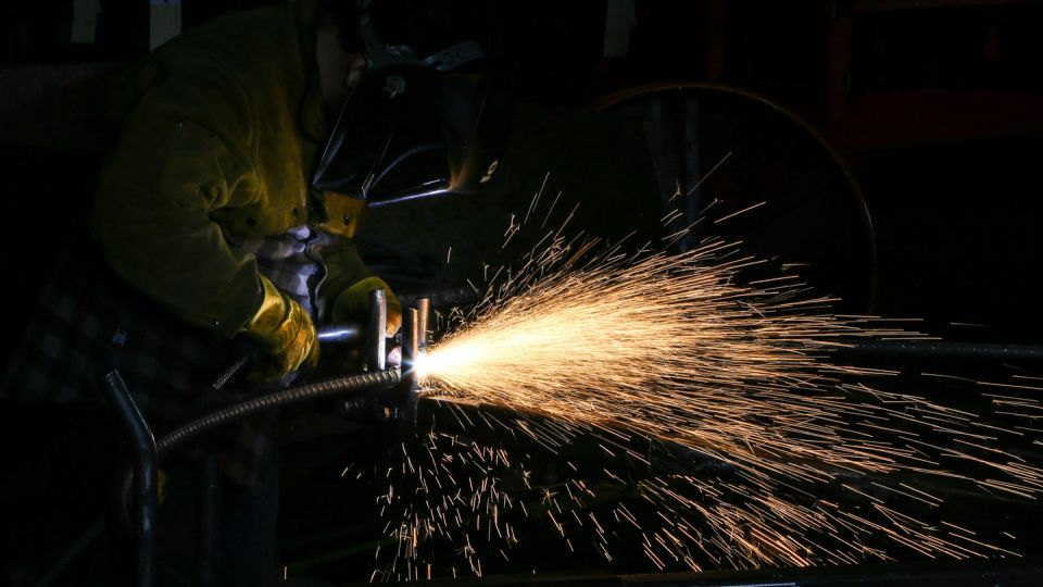A welder in a protective mask and yellow gloves welding metal, sending a bright cascade of sparks across a dark workshop.